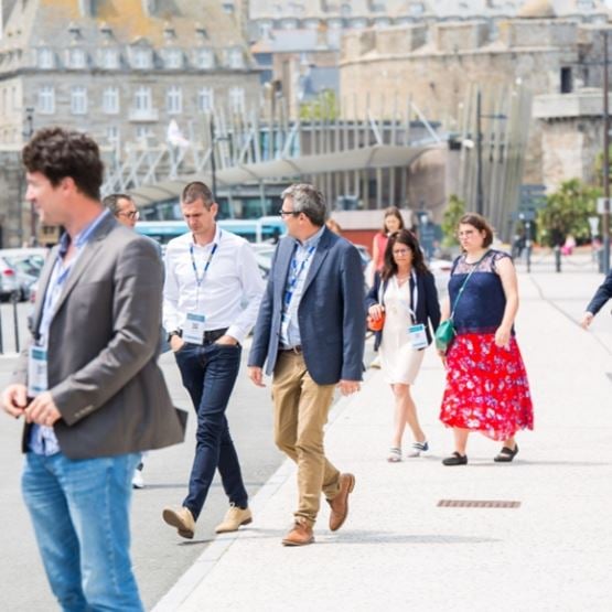 Groupe de personnes marchant sur le trottoir vers le Quai Saint-Malo, devant les murailles fortifiées et les bateaux de Saint-Malo